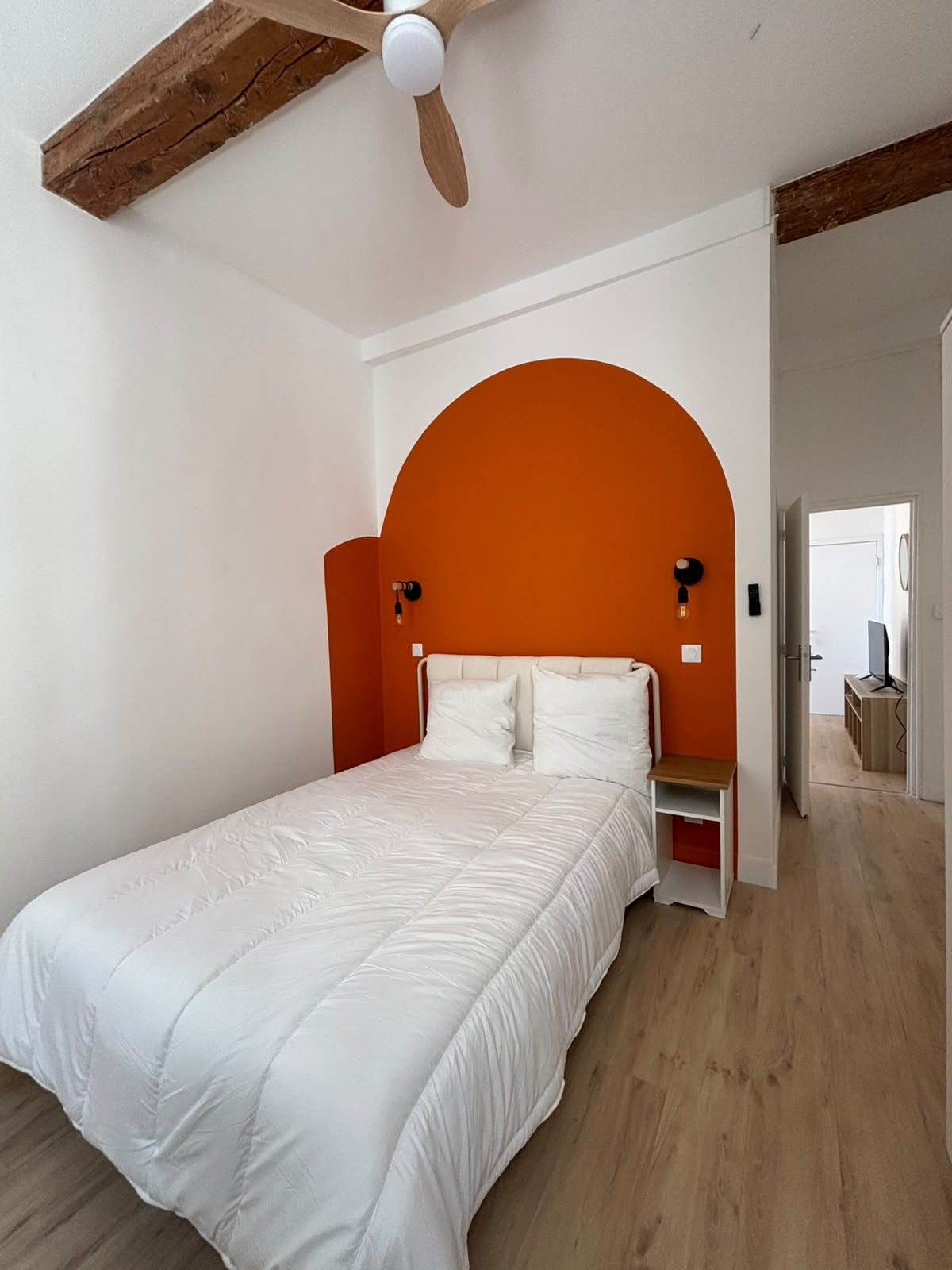 Chambre du T2 rénové à Marseille — tête de lit en arche terracotta, appliques noires et poutres apparentes